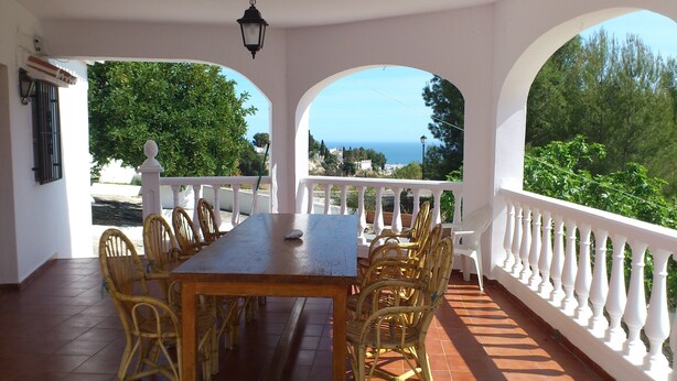 A covered patio with a wooden table and wicker chairs, white balustrades, and a view of the sea.