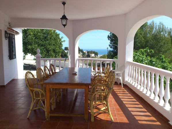 Une terrasse couverte avec une table en bois et des chaises en osier, des balustrades blanches et une vue sur la mer.