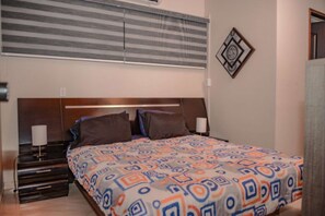Une chambre à coucher comprenant un lit, une table de chevet, une lampe, une fenêtre avec des stores et un élément de décoration murale.