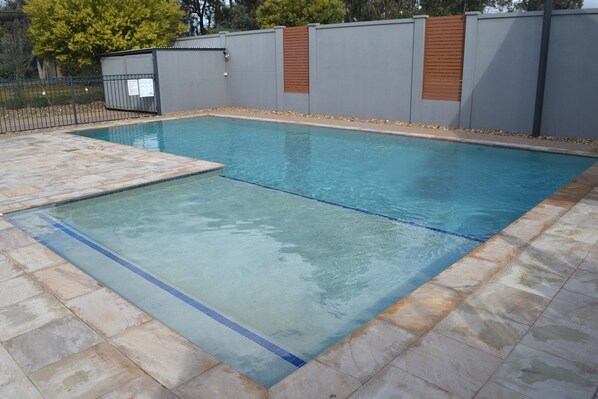A rectangular swimming pool with clear blue water, surrounded by a tiled deck and a grey wall with wooden panels. - Waterview- Stoneleigh at Yarra (Yarrawonga)