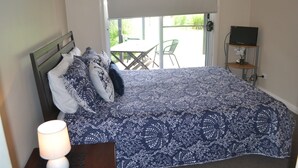 Un dormitorio con cama, mesita de noche con lámpara, televisor sobre un mueble bajo y vistas a un patio exterior con sillas y mesa.