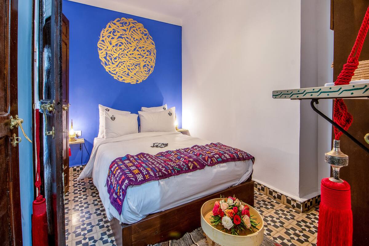 Un dormitorio con una pared azul de acento, una cama de madera, un edredón estampado y una pequeña mesa de madera con un arreglo floral.