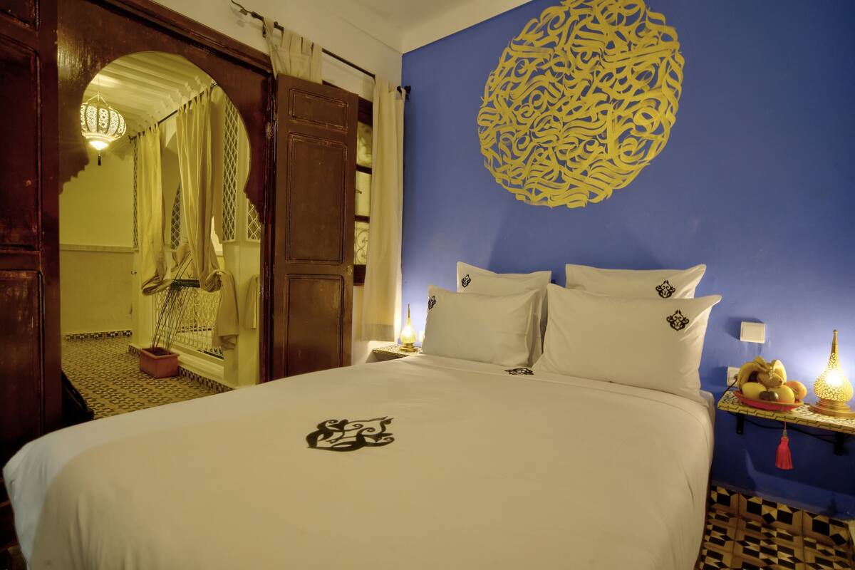 Una habitación de hotel con una cama, un adorno en la pared, una mesita de noche con una lámpara y una ventana con cortinas.