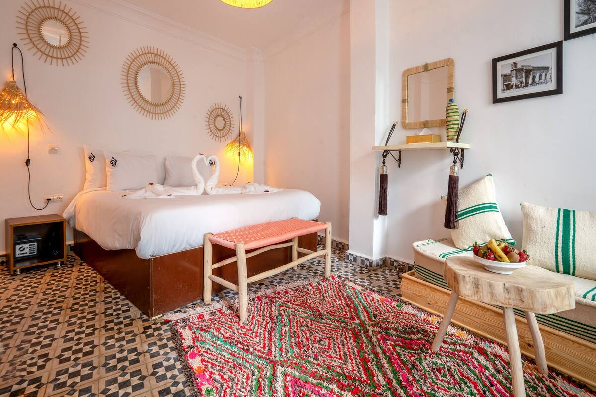 Habitación de hotel con cama, un banco, una mesita con fruta, alfombra estampada y decoración en la pared.