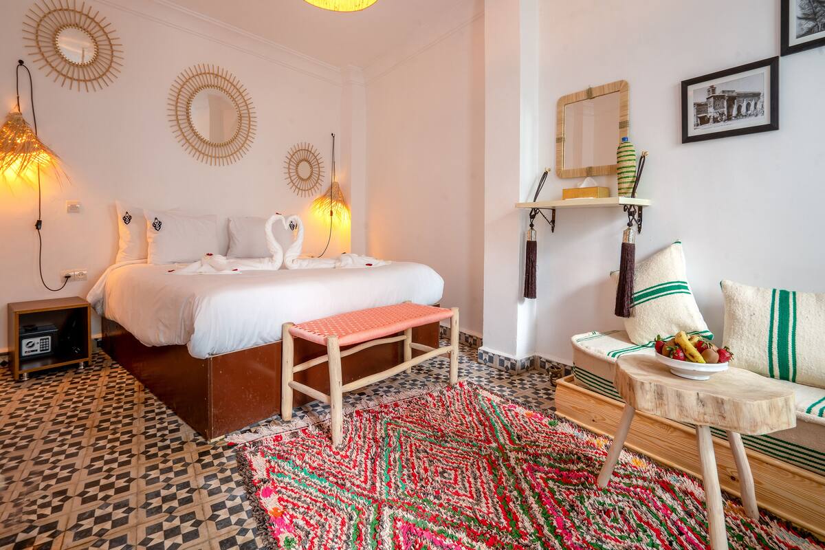 Habitación de hotel con cama, un banco, una mesita con fruta, alfombra estampada y espejos decorativos en la pared.