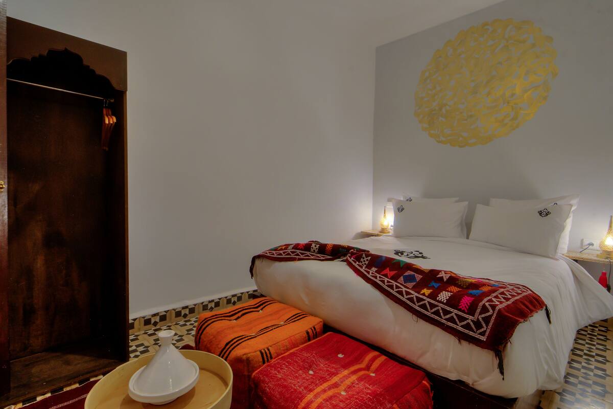 Un dormitorio con cama, almohadas, manta, un adorno en la pared y una mesita de noche con lámpara.