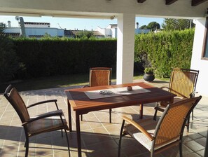 Een terras met een houten tafel en rieten stoelen, omgeven door een witte muur en een haag.