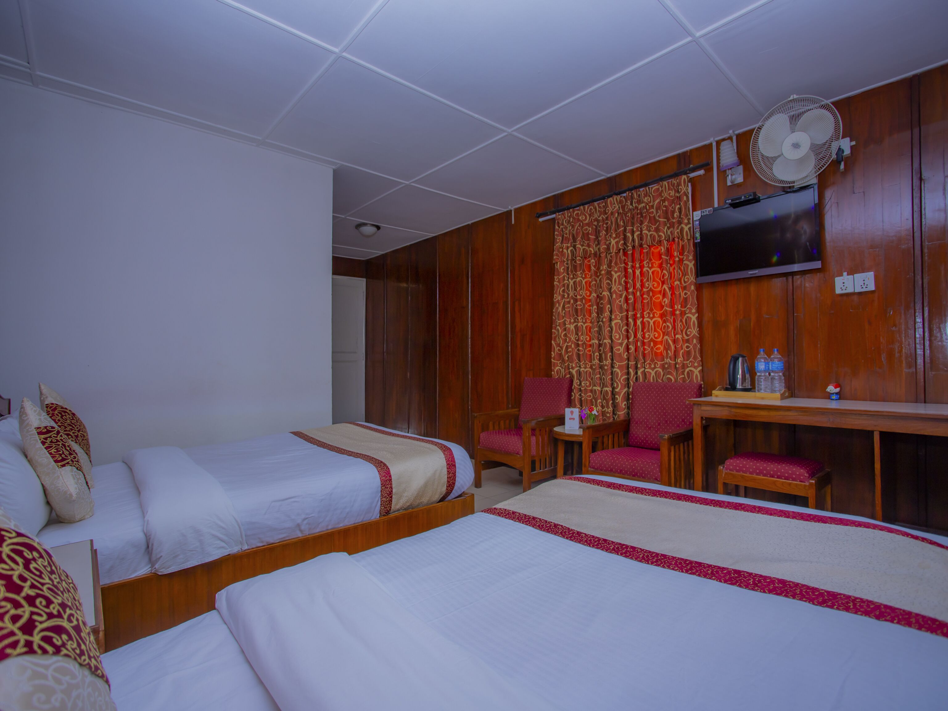 A hotel room with two single beds, a wooden desk with a chair, a TV mounted on the wall, and a ceiling fan.