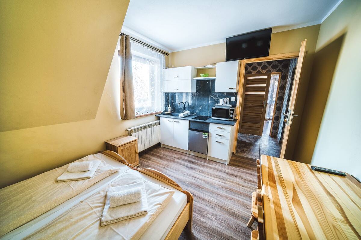 A compact room with a bed, a wooden table, a kitchenette, and a bathroom.