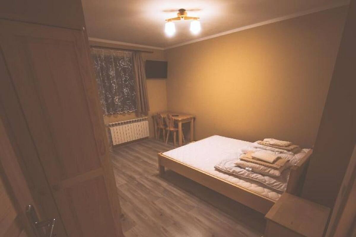 A bedroom with a bed, a desk, and a chair. There is a window with curtains and a radiator on the wall.