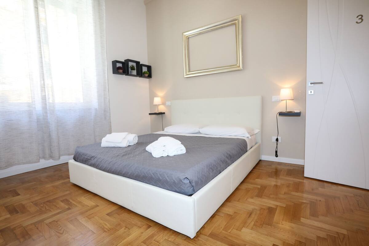 Un lit bien fait, avec du linge de lit blanc et une couvre-lit grise, une table de chevet avec une lampe, un tableau encadré au mur et une porte portant le numéro 3.