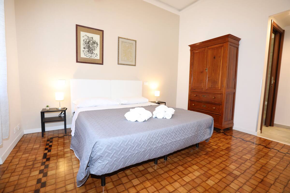 Une chambre à coucher bien rangée, avec un grand lit, deux tables de chevet, une armoire en bois et deux tableaux encadrés accrochés au mur.