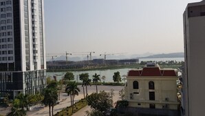 Um edifício alto com design moderno, um rio e guindastes de construção ao fundo, e um edifício tradicional com telhado vermelho em primeiro plano.