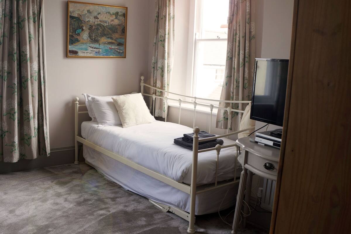 A bedroom with a single bed, a painting on the wall, a window with curtains, and a television on a stand.