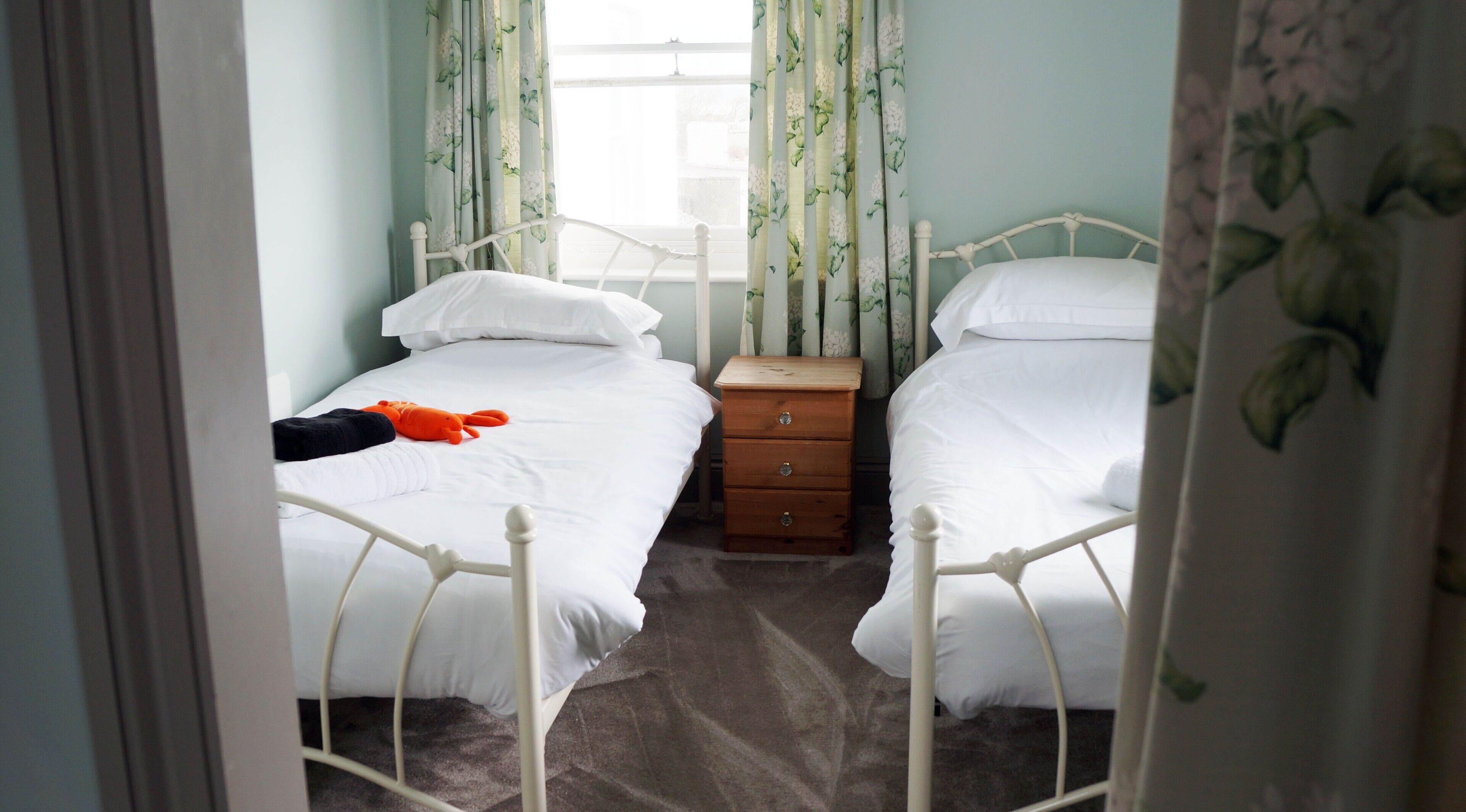 Quarto com duas camas de solteiro, mesa de cabeceira de madeira e cortinas florais.