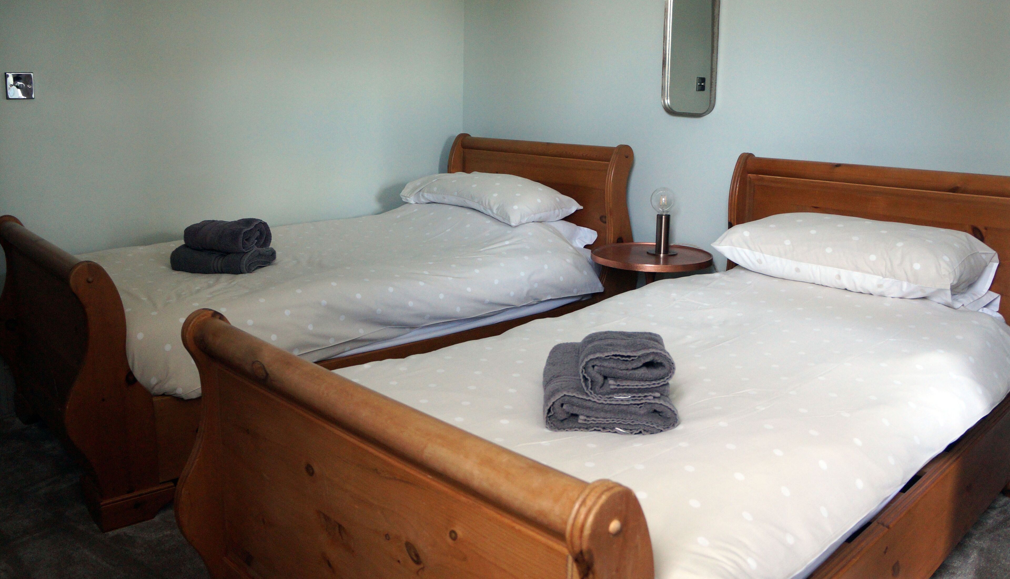 A room with two wooden beds, each with a pillow and a rolled towel. There is a small table with a lamp between the beds.