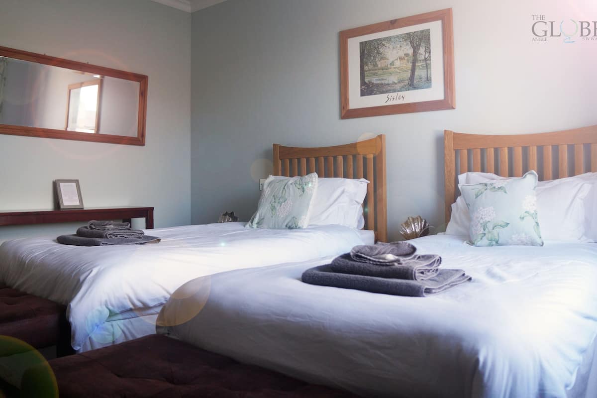 A hotel room with two single beds, each with a wooden headboard, white bedding, and decorative pillows. A framed picture hangs on the wall above the beds.
