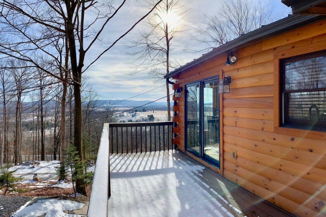 A wooden cabin with a deck, large windows, and a view of a snowy landscape.