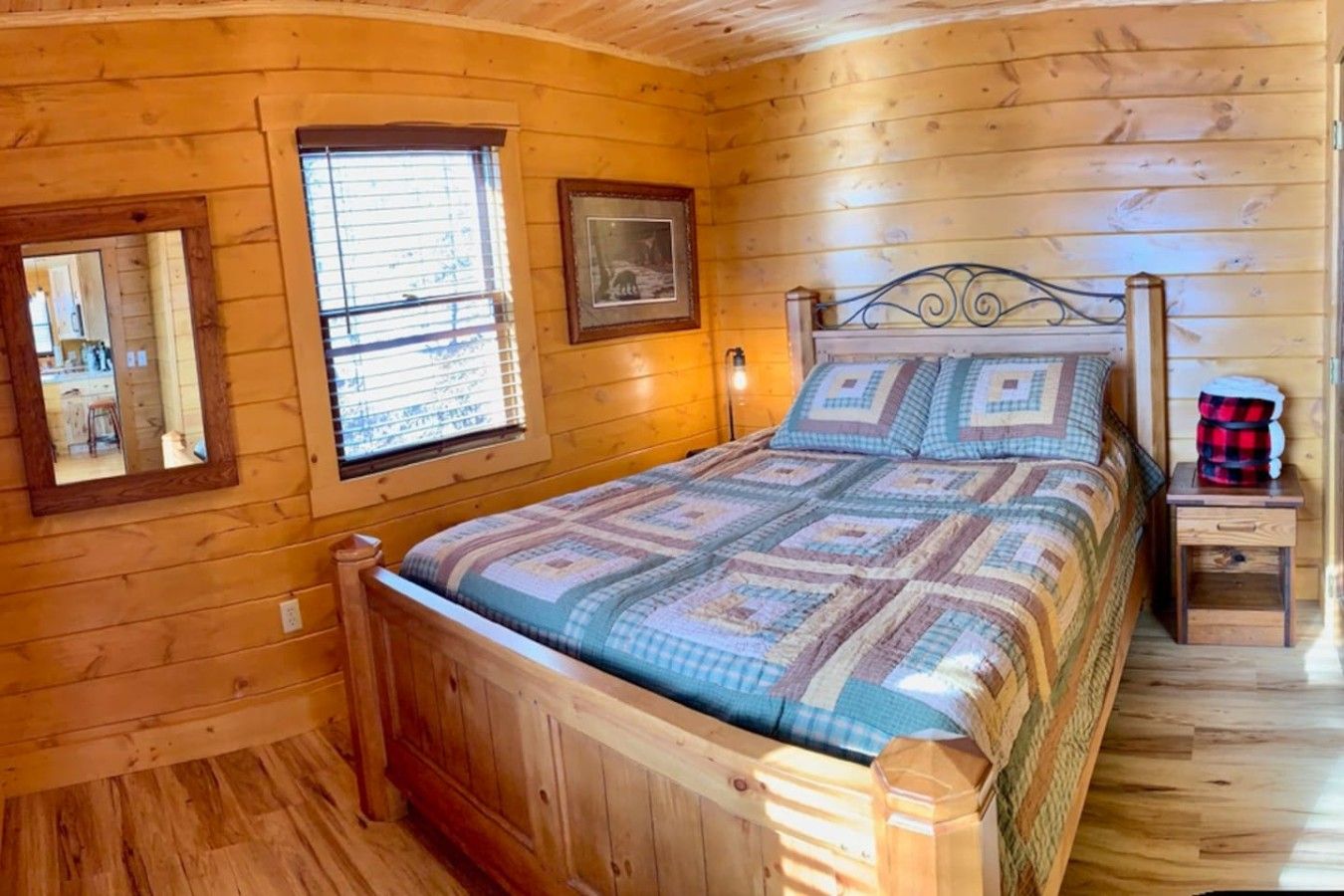 A wooden cabin bedroom with a bed, a window, a framed picture, and a nightstand.