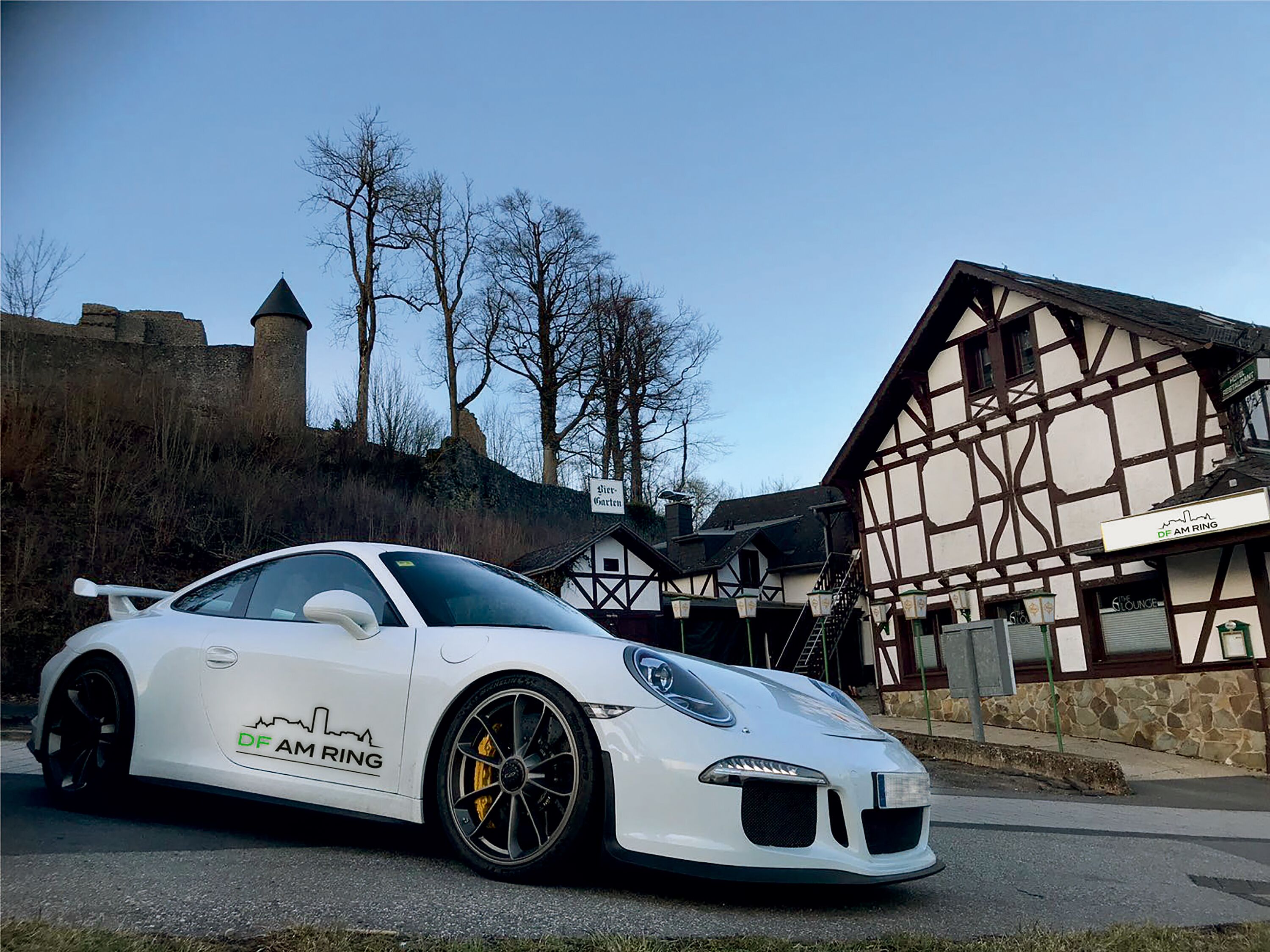A white sports car parked in front of a traditional half-timbered building with a sign that reads "DF AM RING".