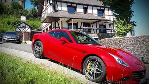 A red sports car parked in front of a traditional half-timbered house with a stone wall and a small wooden structure to the left. -  DF am Ring Nürburg (Nuerburg)
