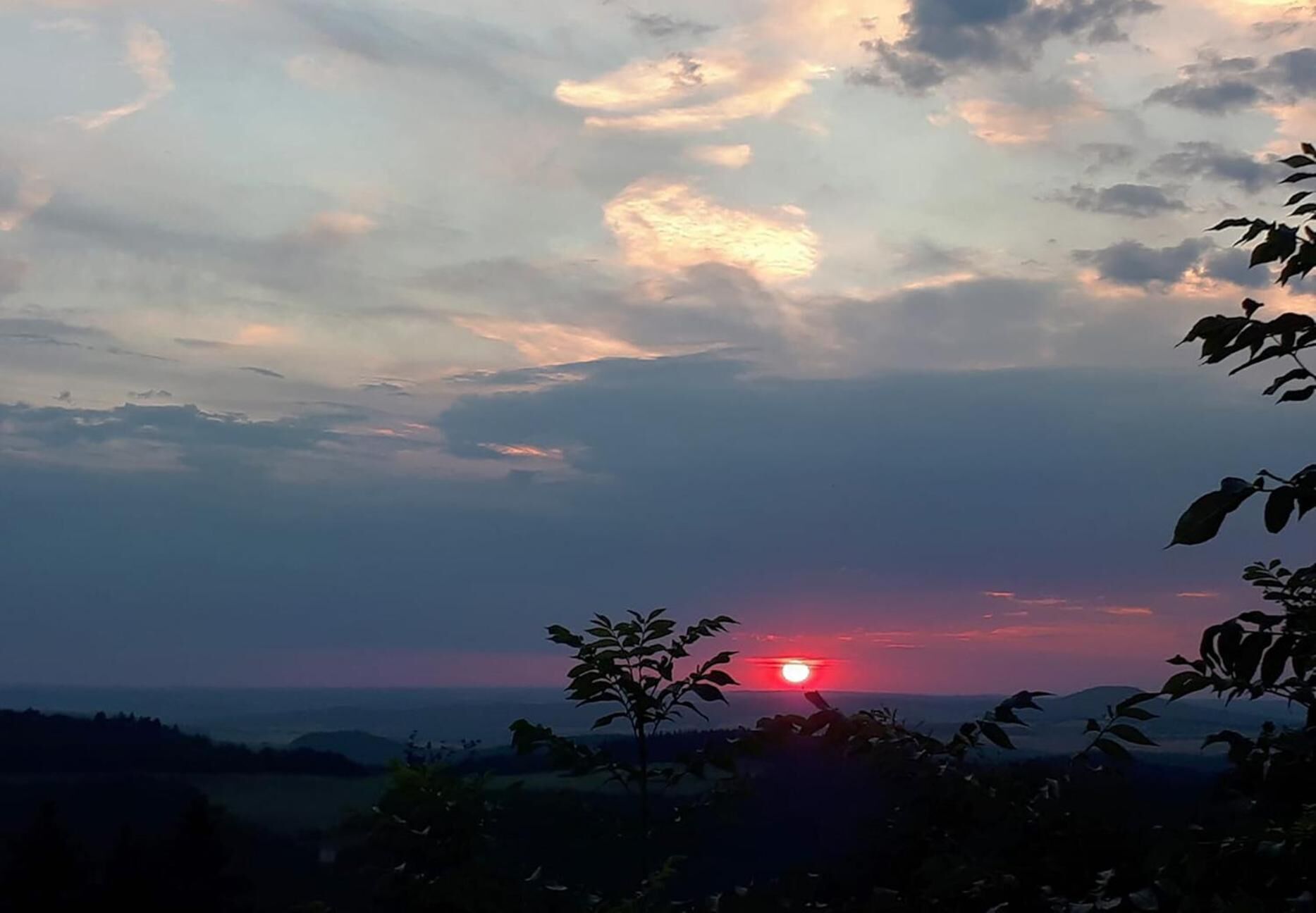 A sunset over a landscape with silhouettes of trees and distant hills.