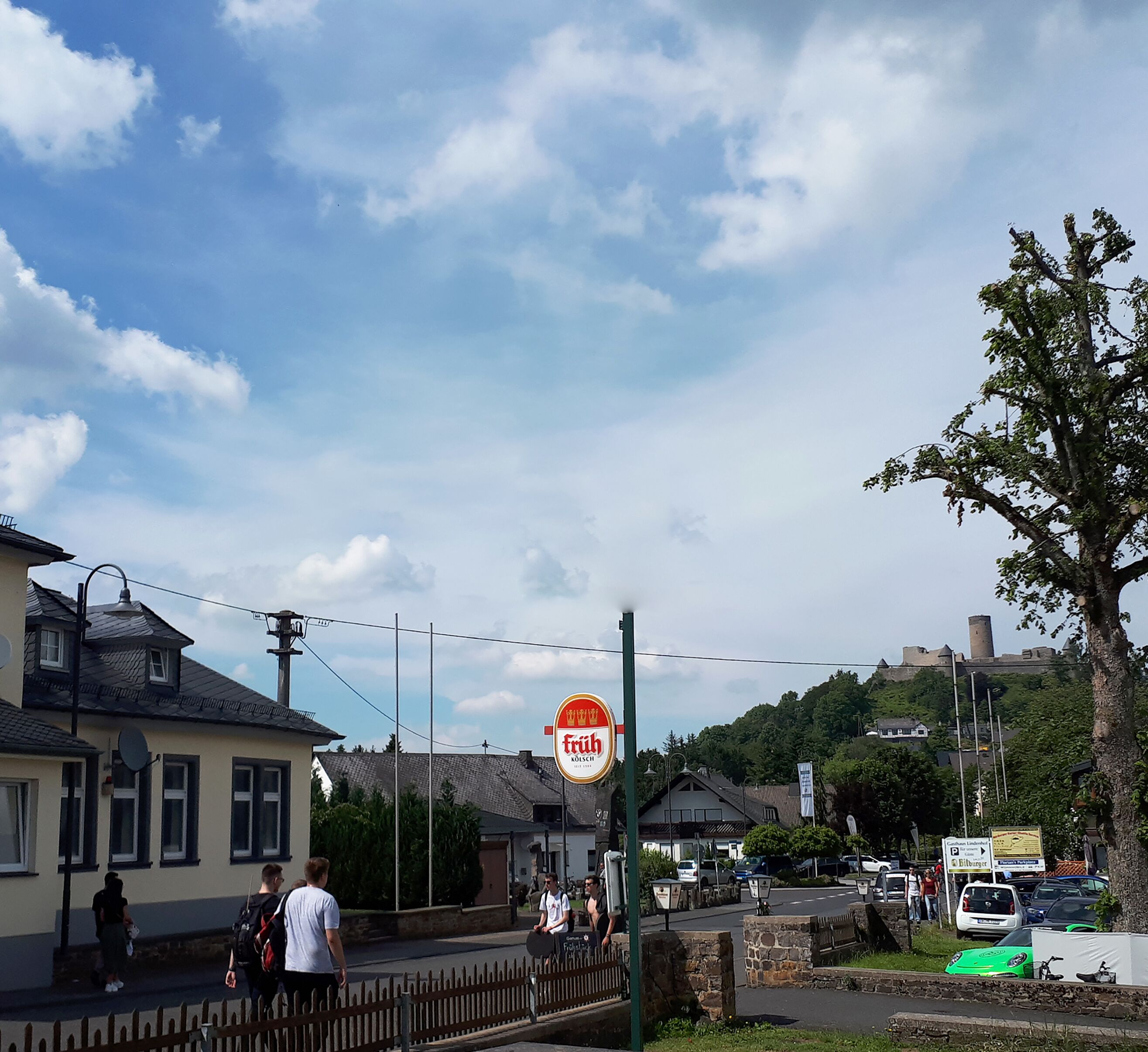 Straßenansicht mit einem Gebäude, an dem ein Schild für 'Früh' hängt, und einer Burg im Hintergrund.