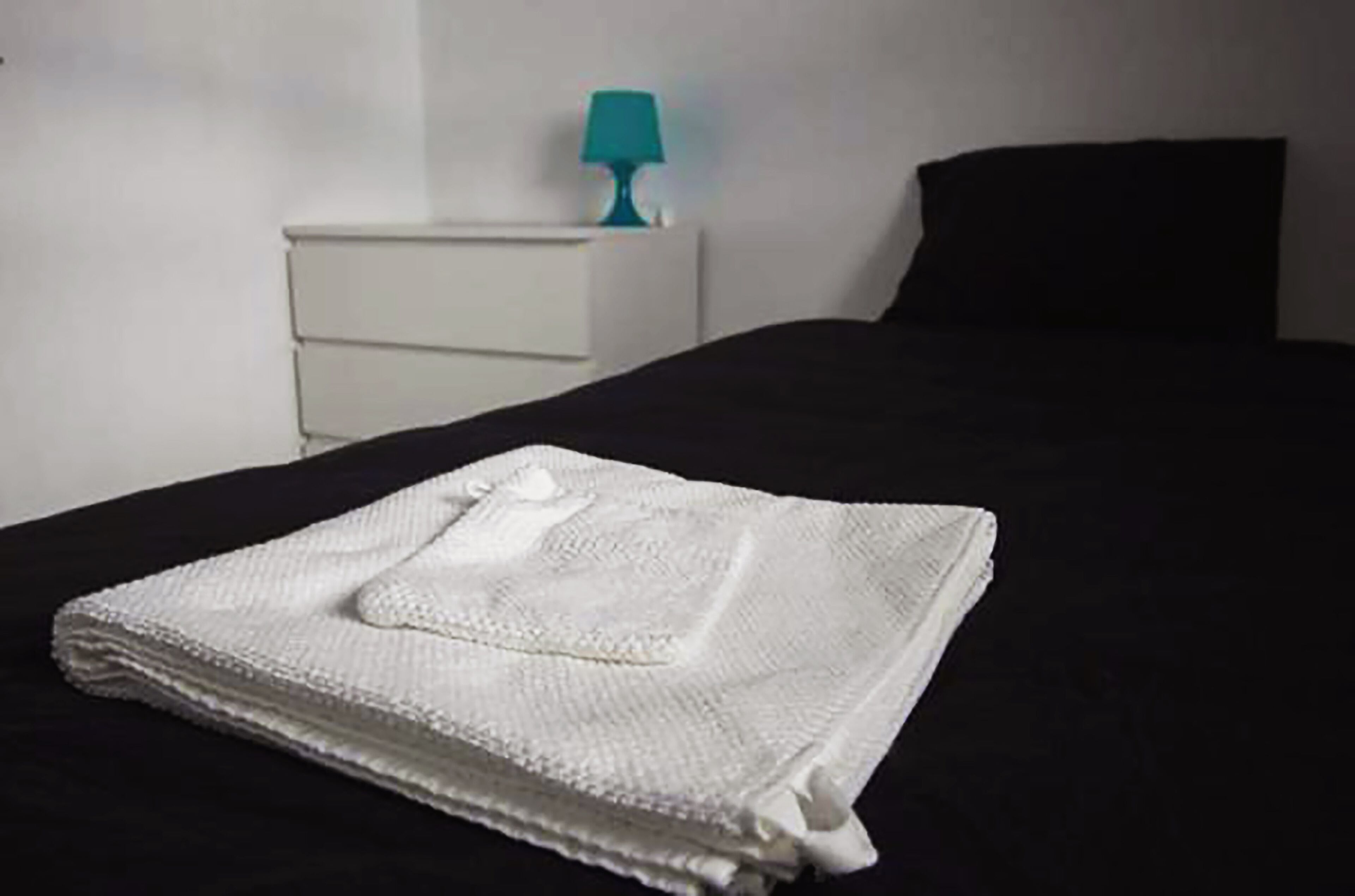 Un lit bien fait, recouvert d’une couverture blanche, avec un oreiller noir et une table de chevet blanche sur laquelle se trouve une lampe turquoise.