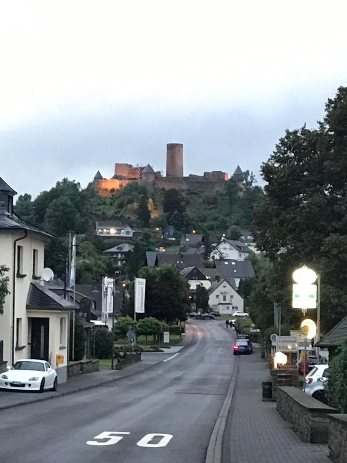 A street leading to a castle-like structure with a tower, surrounded by residential buildings and a paved road marked with a speed limit sign.
