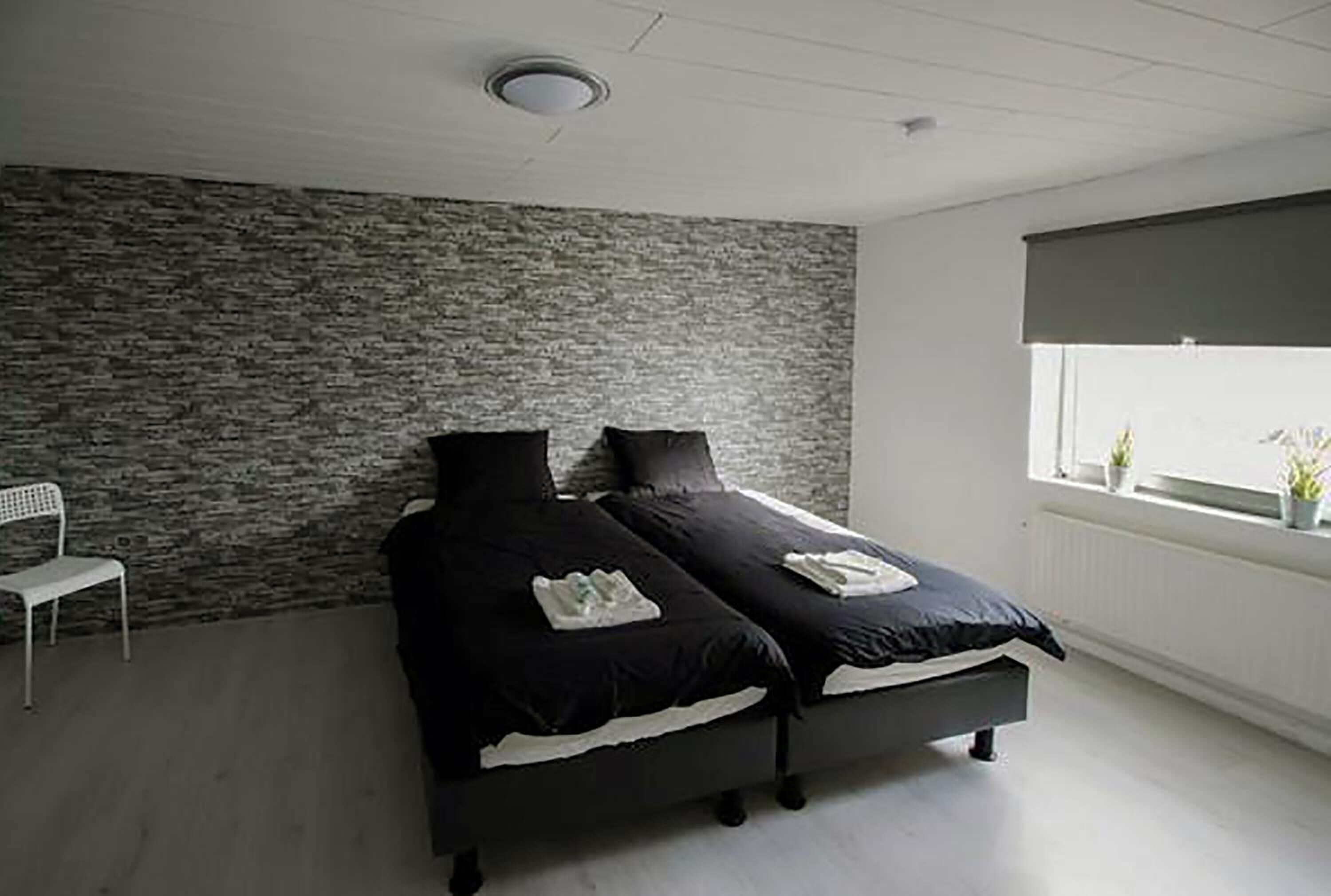 A bedroom with two beds, a white chair, a stone-textured wall, and a window with plants.
