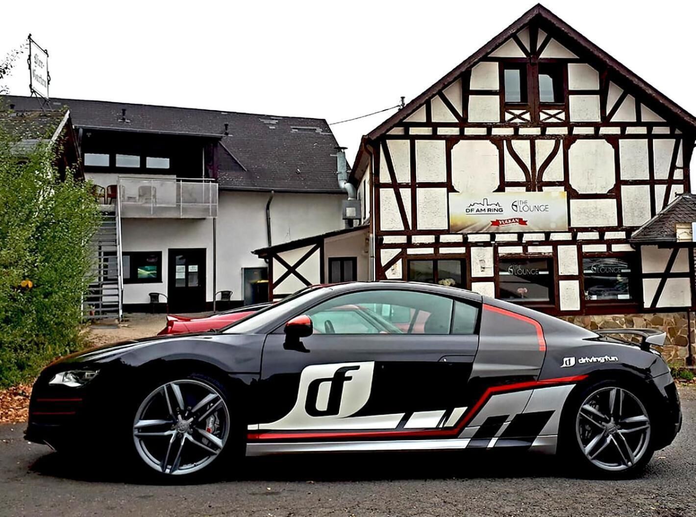 Une voiture de sport noire est garée devant un bâtiment traditionnel à colombages, sur lequel on peut lire l’inscription « The Lounge ».