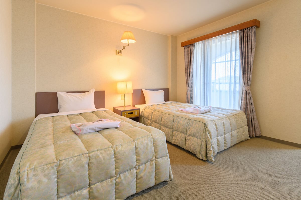 A hotel room with two beds, a nightstand, a lamp, and a window with curtains.