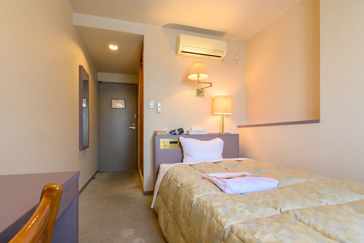A hotel room with a bed, a desk, a chair, an air conditioning unit, and a lamp.