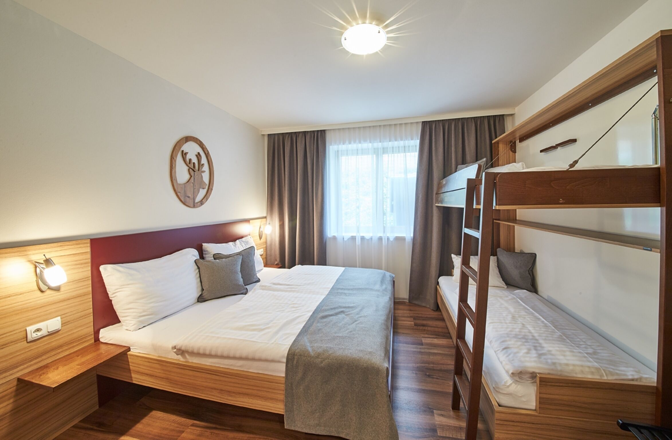 A hotel room with a large bed, a bunk bed, wooden flooring, a wall-mounted light, a window with curtains, and a wall decoration.