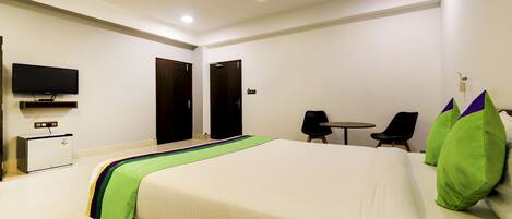 Ein Hotelzimmer mit einem Bett, einem an der Wand befestigten Fernseher, einem kleinen Tisch, zwei Stühlen und einem Deckenventilator.