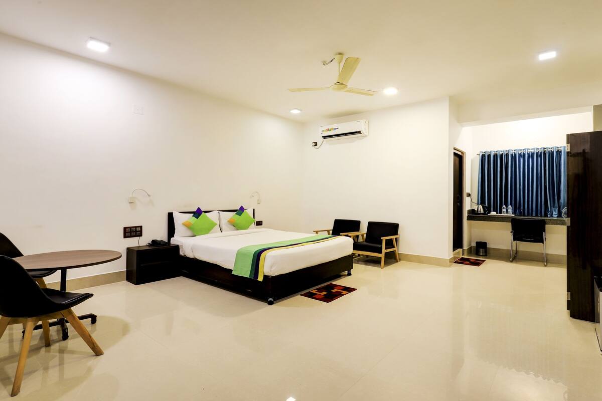 A modern hotel room with a bed, desk, chairs, and a refrigerator.