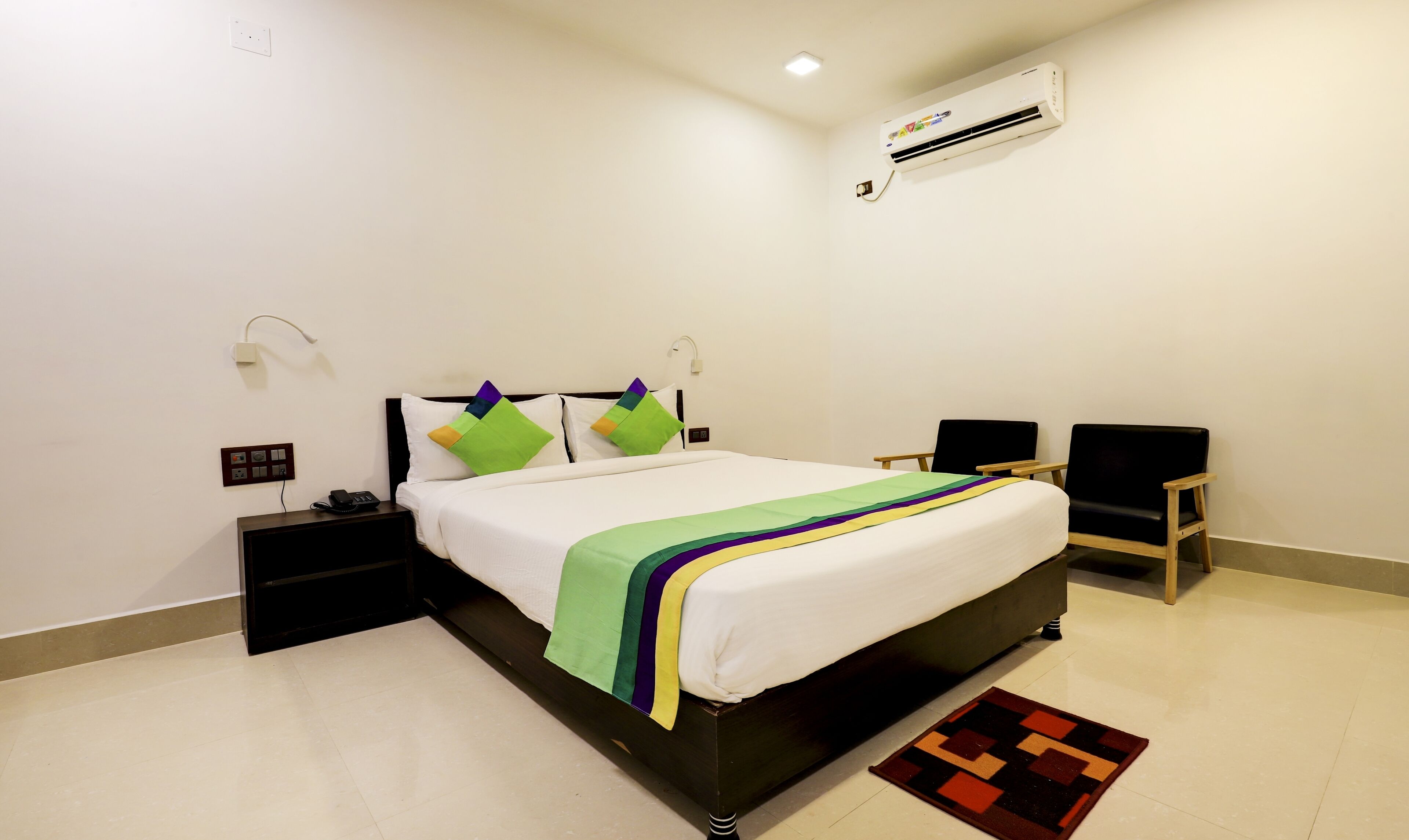 A neatly arranged hotel room with a bed, bedside tables, a chair, and an air conditioning unit.