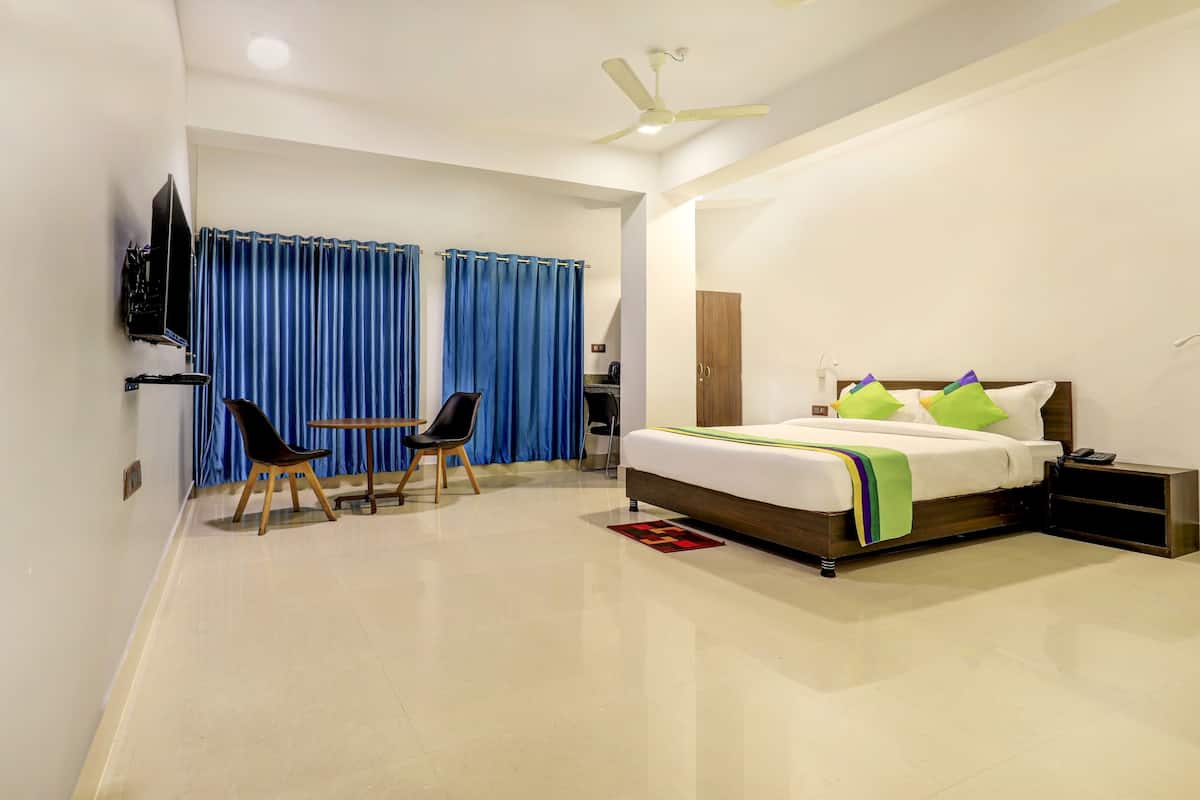 A modern hotel room with a bed, a desk, two chairs, a TV, and a ceiling fan.