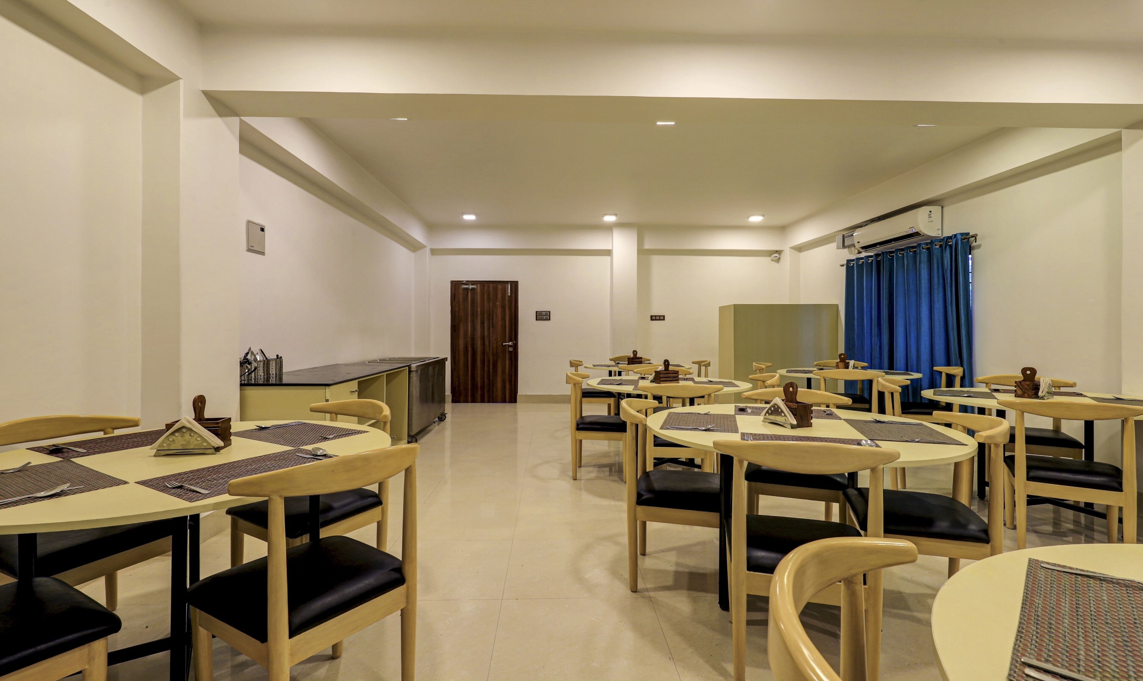 A hotel dining area with multiple tables and chairs, a kitchen area, and a blue curtain.