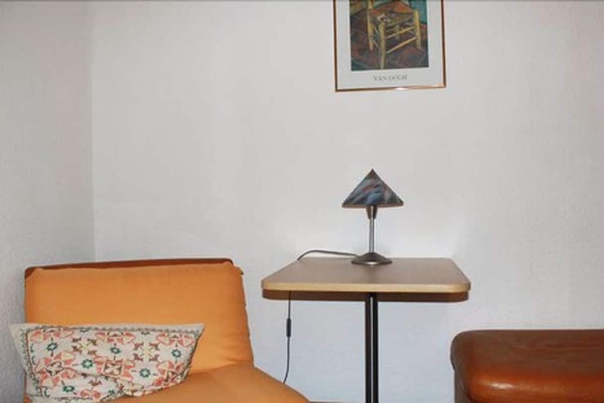 A room with a small table, a cushion, and a framed picture on the wall.