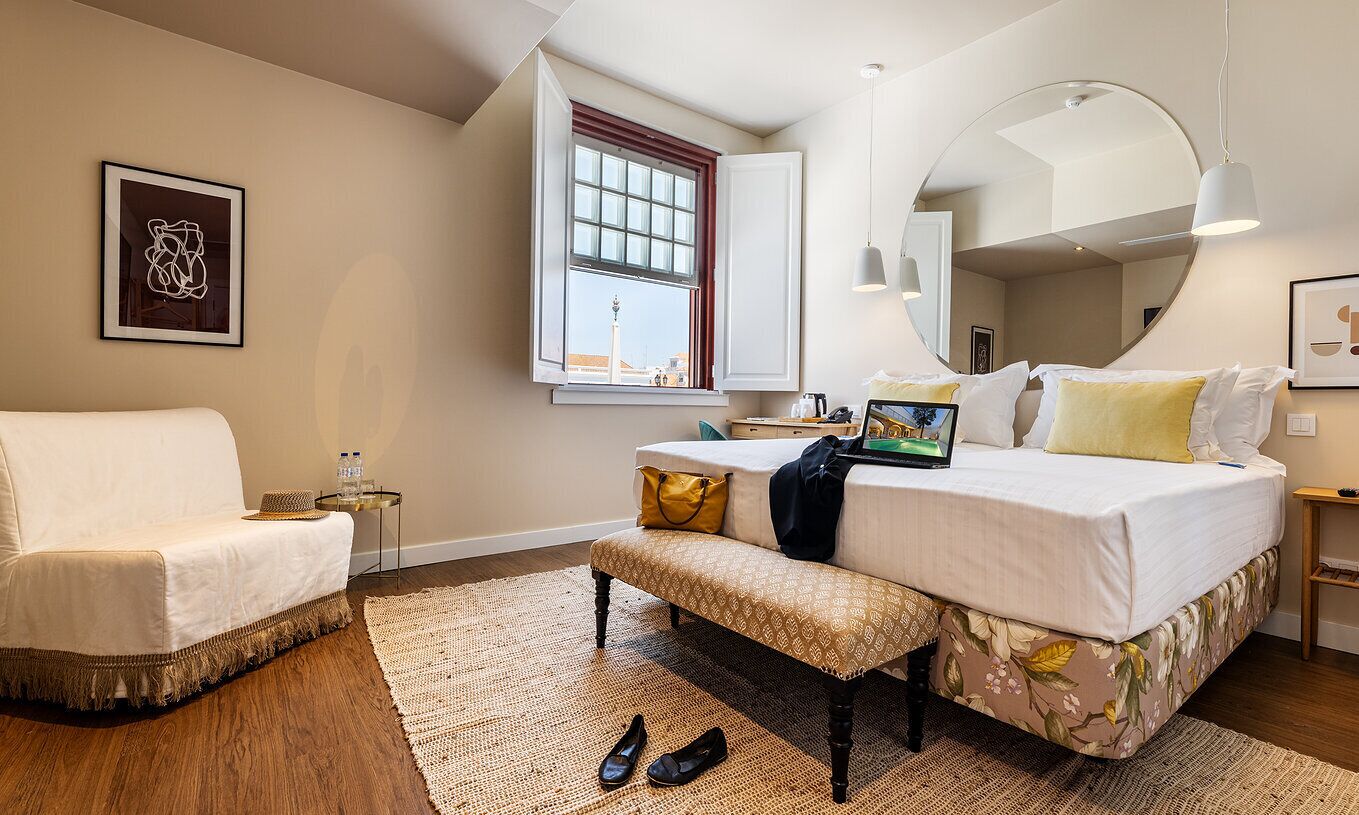 A modern bedroom with a large bed, a bench, a small table, a window with a view, and a mirror.