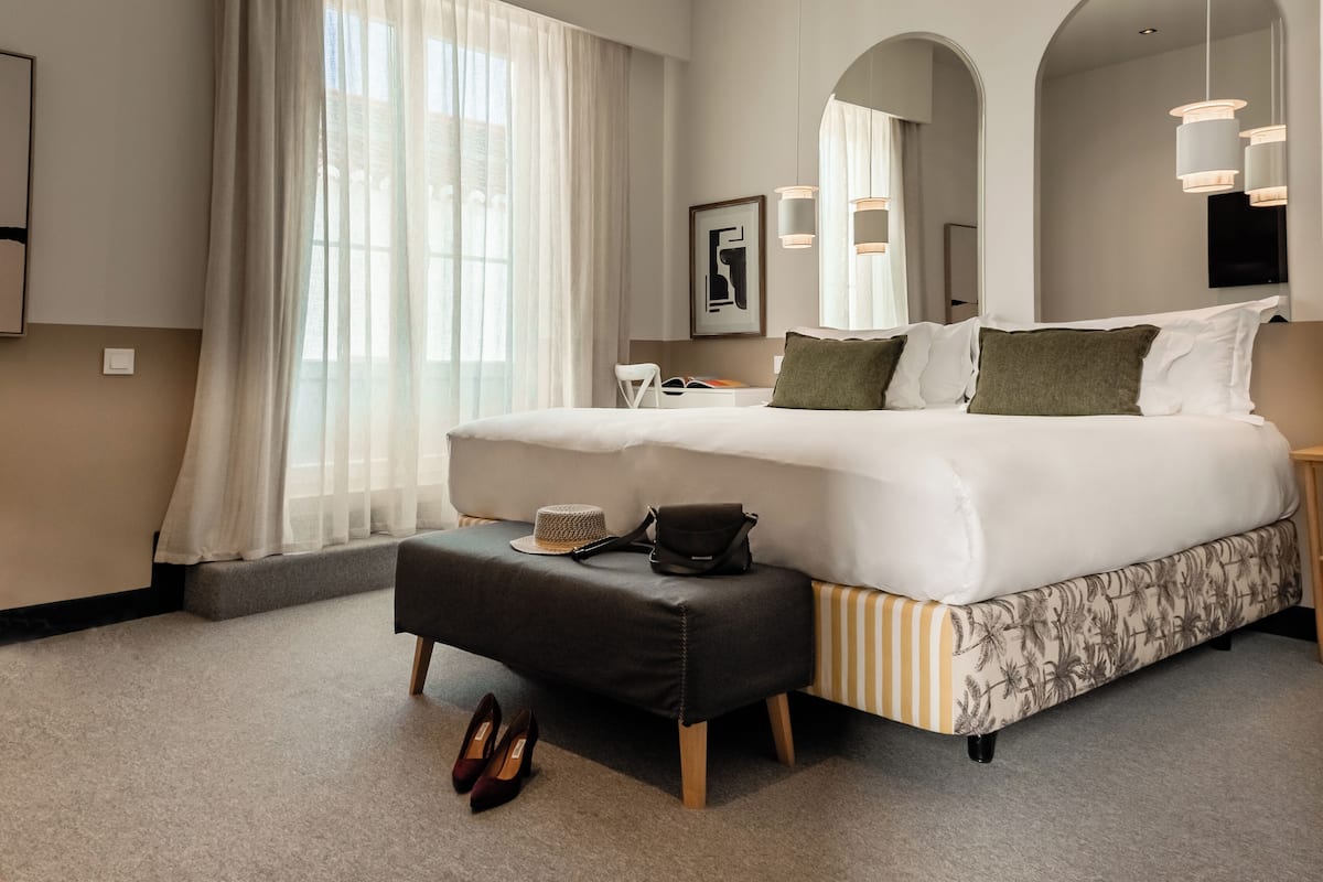 A modern hotel bedroom with a large bed, a bench, a bedside table, and a window with curtains.