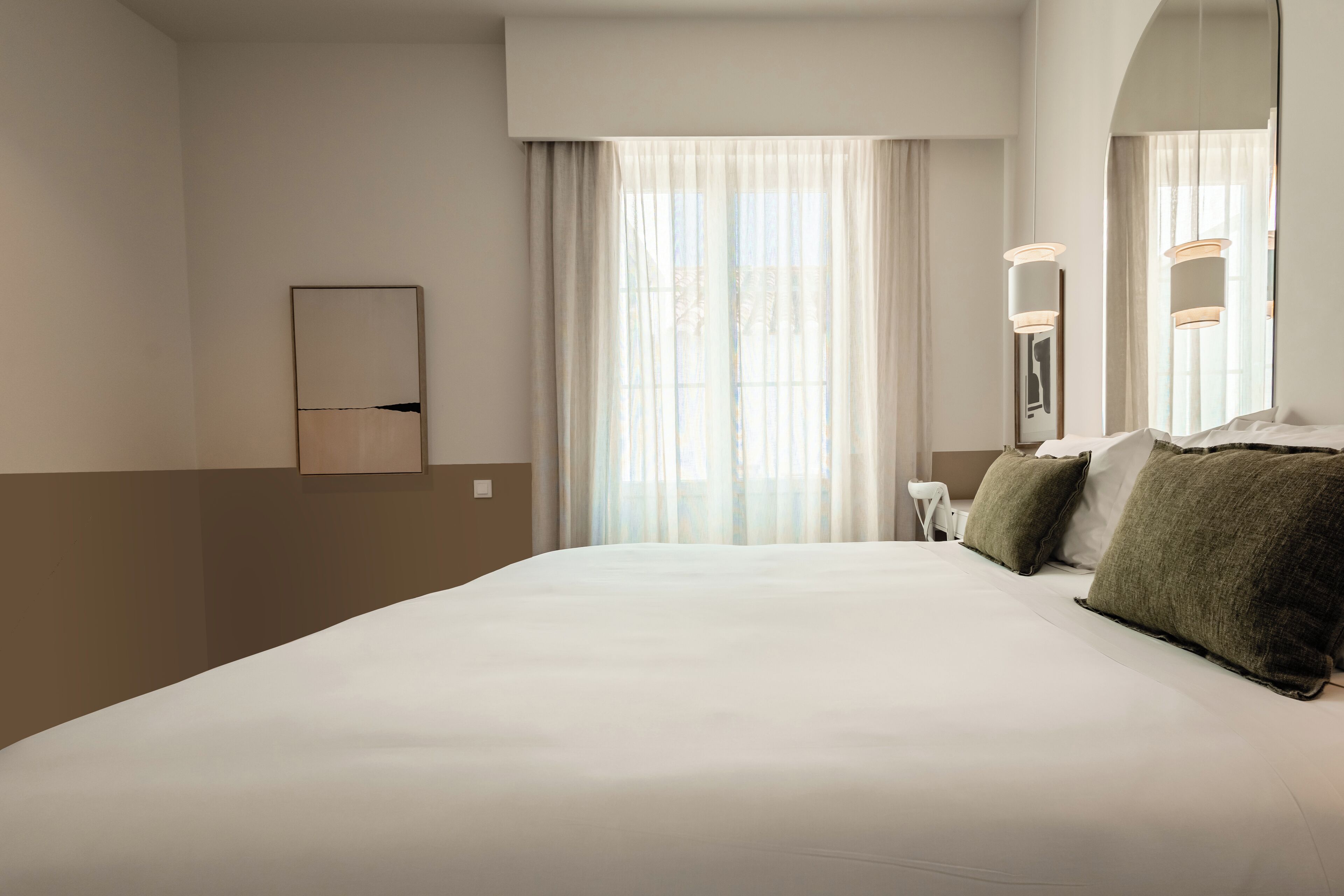 Habitación de hotel con una cama grande, una mesita de noche, una silla y una ventana con cortinas traslúcidas.