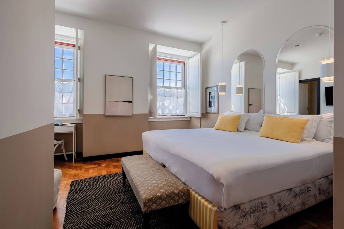A hotel room with a large bed, a desk, and a chair. There are windows with curtains, a mirror, and a television.