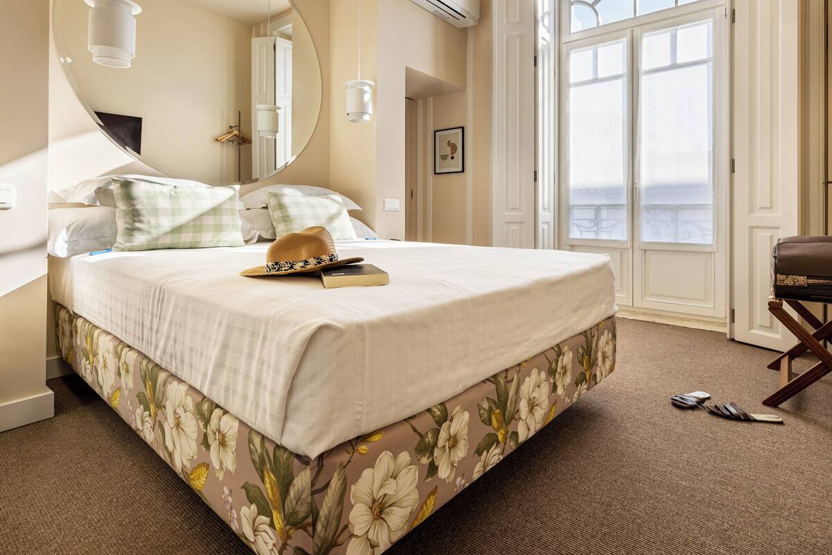 A neatly made bed with a floral bedspread, a round mirror, and a view of the outdoors through a large window.
