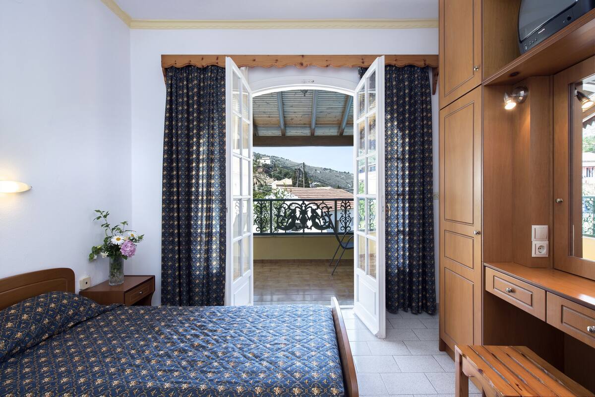 A bedroom with a bed, a nightstand with a vase of flowers, a wooden desk, and a balcony with a view of the hills.