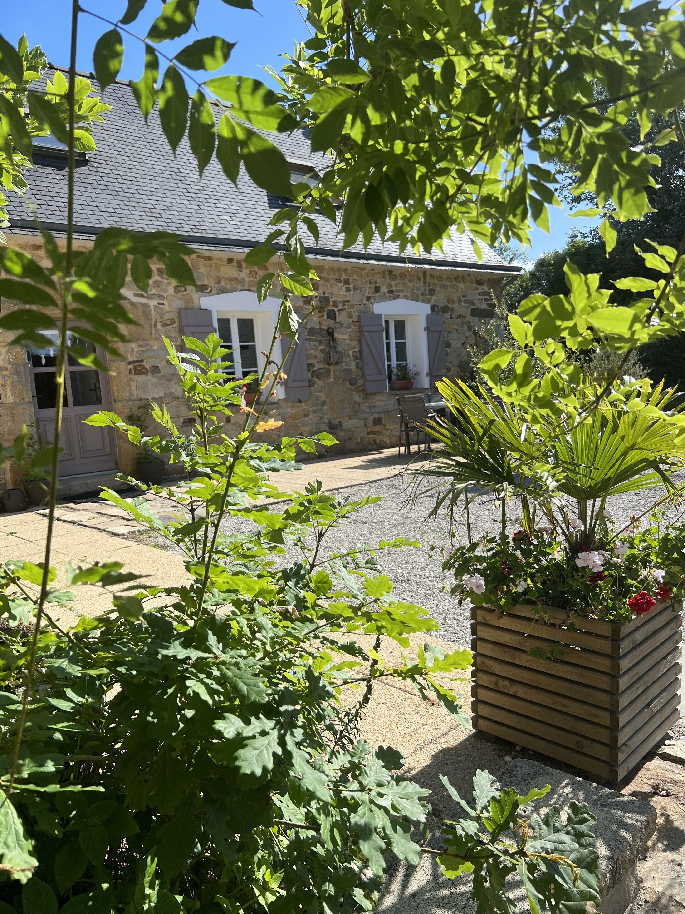 A stone building with a gravel courtyard, surrounded by greenery and a wooden planter with flowers.