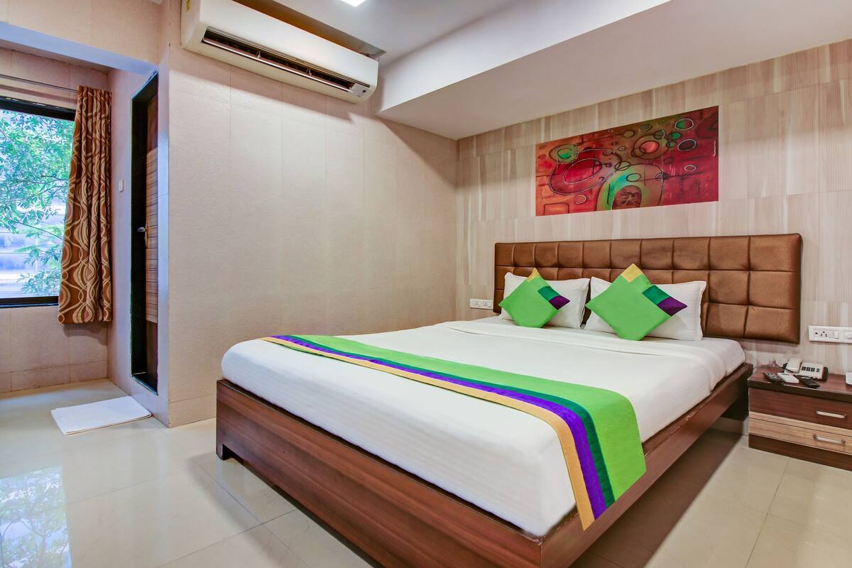 A hotel room with a large bed, a headboard, a painting on the wall, a nightstand, and a window with curtains.