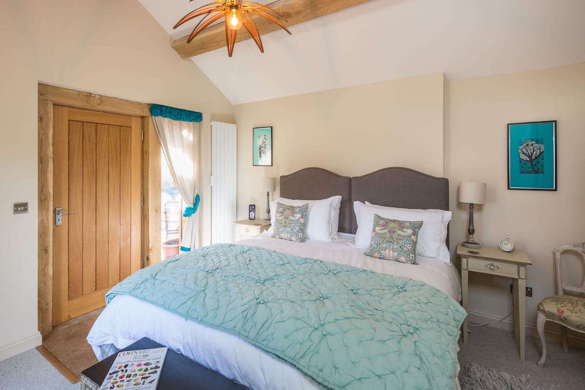 A bedroom with a large bed, wooden beams, a hanging light fixture, a nightstand, a chair, and a dresser.