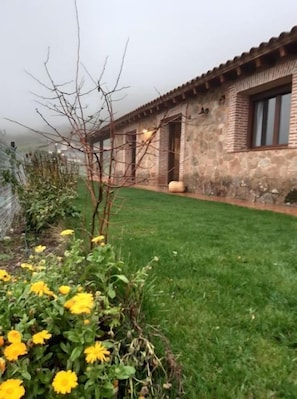 A stone building with a tiled roof, a grassy lawn, and yellow flowers in the foreground. - Beautiful house surrounded by meadows and oaks, with private pool and excellent views of the valley (El Raso -Candeleda)
